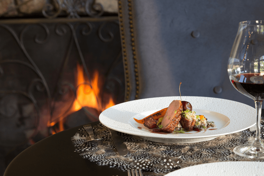 Gourmet duck breast entrée served with red wine in front of a glowing fireplace